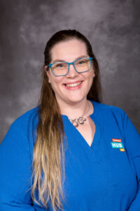 A photo of Marcy LaCroix, a woman with long brown hair who is wearing glasses and a blue shirt with the Disability Hub MN logo on it.