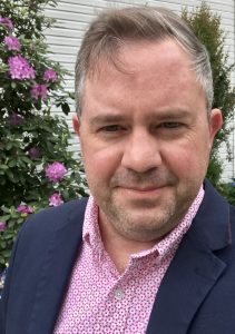 Bill McCollam, a white man with grey hair and stubble wearing a pink shirt and blazer